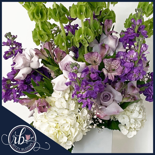 Fresh flower arrangement with lavender roses, purple stock, lavender alstroemerias, Bells of Ireland, and white hydrangeas in a white vase