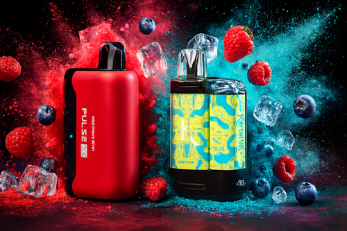 The photo shows two vape devices surrounded by dynamic splashes of color, fruit, and ice cubes. On the left is a red and black device labeled &ldquo;PULSE&rdquo; with the flavor &ldquo;WILD CHERRY BATCH&rdquo; printed vertically. On the right is a more elaborate design featuring yellow and blue patterns, marked &ldquo;ICE POP&rdquo; and &ldquo;Kartel 9K.&rdquo; Around them are raspberries, blueberries, and ice cubes, while the background bursts with red and blue powder effects, emphasizing the fruity, refreshing flavors and bold branding of the products.