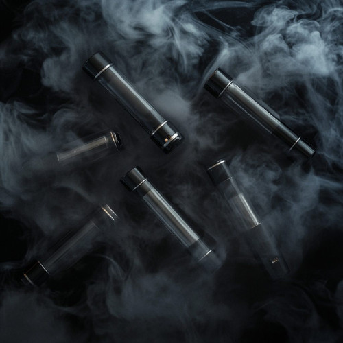 The image shows several sleek cylindrical objects, likely vape pens or electronic cigarettes, arranged artistically amidst swirling clouds of smoke. These vape pens feature a metallic black design, exuding a modern and stylish appeal. The dramatic smoke surrounding them creates a sense of mystery and highlights the aesthetic allure of the vaping devices. This composition effectively draws attention to the clean lines and sophisticated design of the products.