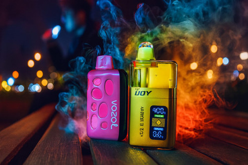 Two vibrant disposable vapes, one pink and one gold, sit on a wooden surface against a backdrop of colorful smoke and blurry city lights. The pink device features a unique textured pattern, while the gold device displays a digital screen and the "IJOY" brand name. The overall scene is atmospheric and cinematic, highlighting the sleek designs of the electronic devices through dramatic lighting.