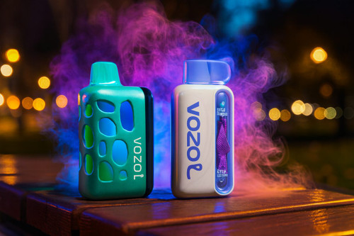 Two VOZOL brand disposable vapes are displayed on a wooden surface against a dark background with golden bokeh city lights. The device on the left is teal with a unique honeycomb-style cutout exterior, while the one on the right is white with a transparent blue mouthpiece and side panel. Swirls of vibrant pink and blue smoke surround the devices, creating a colorful and atmospheric nighttime scene.