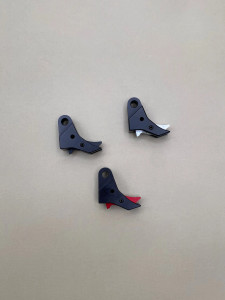 Top view of three Kineti‑Tech style 2 flat‑face trigger shoes arranged in a triangular layout on a smooth tan background. Each trigger shoe is black with a different safety insert color visible: one black, one silver, and one red. All three components are showing their shape, machining details, and color variations clearly.

Three black Kineti‑Tech style 2 flat‑face trigger shoes shown in black, with visible safety inserts in black, silver, and red, arranged on a neutral tan background.