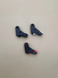 Top view of three Kineti‑Tech style 3 flat‑face trigger shoes arranged in a triangular layout on a smooth tan background. Each trigger shoe is black with a different safety insert color visible: one black, one silver, and one red. All three components are showing their shape, machining details, and color variations clearly.

Three black Kineti‑Tech style 3 flat‑face trigger shoes shown in black, with visible safety inserts in black, silver, and red, arranged on a neutral tan background.