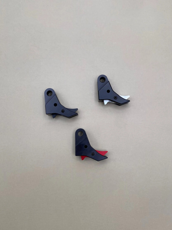 Top view of three Kineti‑Tech style 2 flat‑face trigger shoes arranged in a triangular layout on a smooth tan background. Each trigger shoe is black with a different safety insert color visible: one black, one silver, and one red. All three components are showing their shape, machining details, and color variations clearly.

Three black Kineti‑Tech style 2 flat‑face trigger shoes shown in black, with visible safety inserts in black, silver, and red, arranged on a neutral tan background.