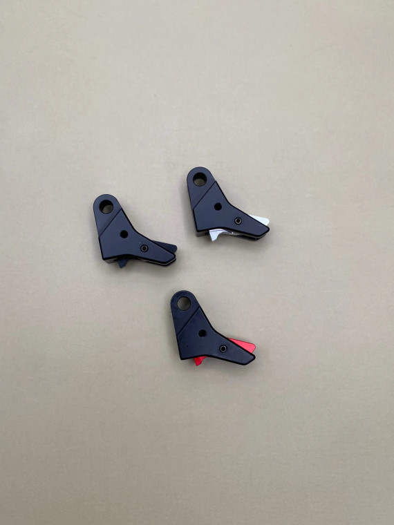 Top view of three Kineti‑Tech style 4 flat‑face trigger shoes arranged in a triangular layout on a smooth tan background. Each trigger shoe is black with a different safety insert color visible: one black, one silver, and one red. All three components are showing their shape, machining details, and color variations clearly.

Three black Kineti‑Tech style 4 flat‑face trigger shoes shown in black, with visible safety inserts in black, silver, and red, arranged on a neutral tan background.