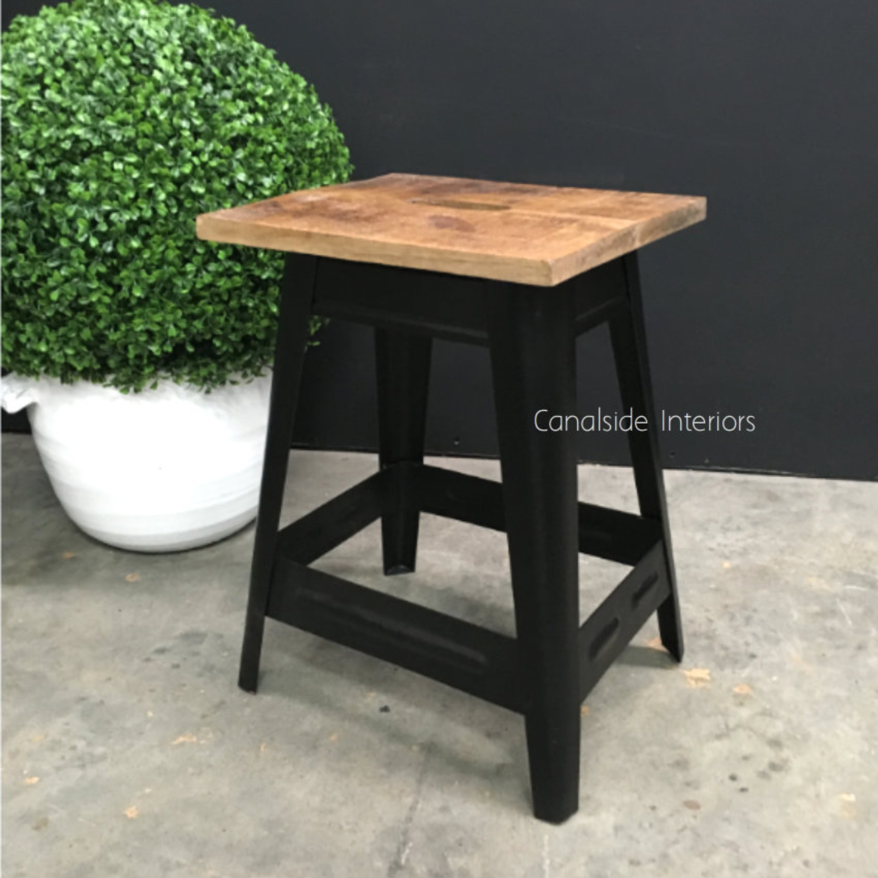 The Garden Drive Stool by Canalside Interiors showcases a distressed black finish with a natural wood top, a versatile piece for a Hamptons-inspired bedroom or a coastal-themed sitting area
