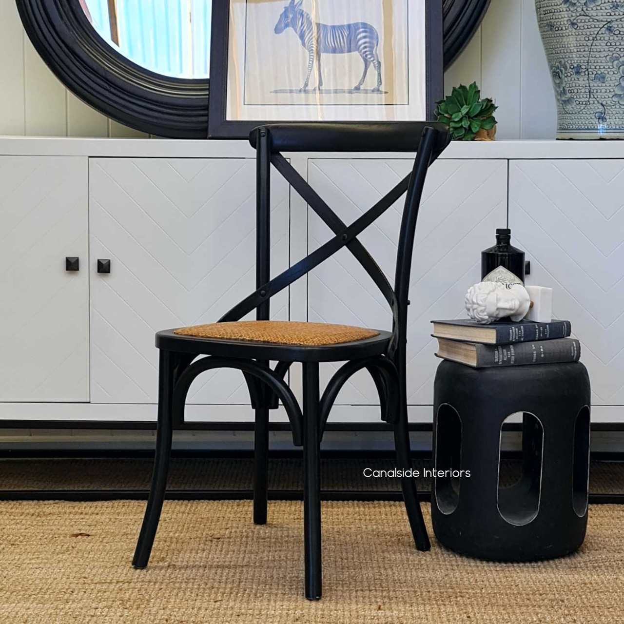 The natural rattan seat juxtaposed with the distressed black frame of Canalside Interiors' Cross Back Chair offers an inviting addition to any living room decor.
