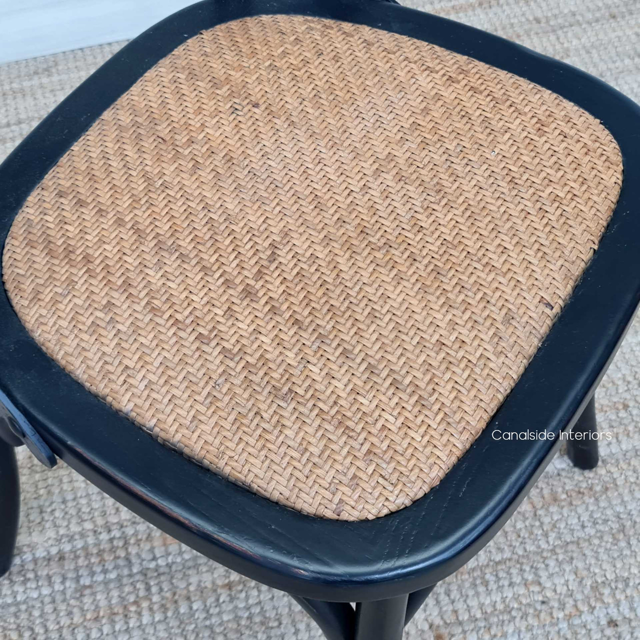 Featuring a durable rattan seat, the Cross Back Chair from Canalside Interiors in distressed black adds a touch of elegance to a living room with its coastal and classical design