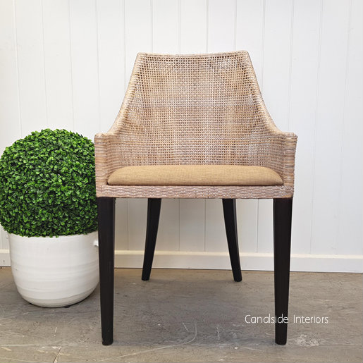 The chair positioned against a crisp white wall, with the natural light accentuating the texture of the rattan and the softness of the upholstered seat.