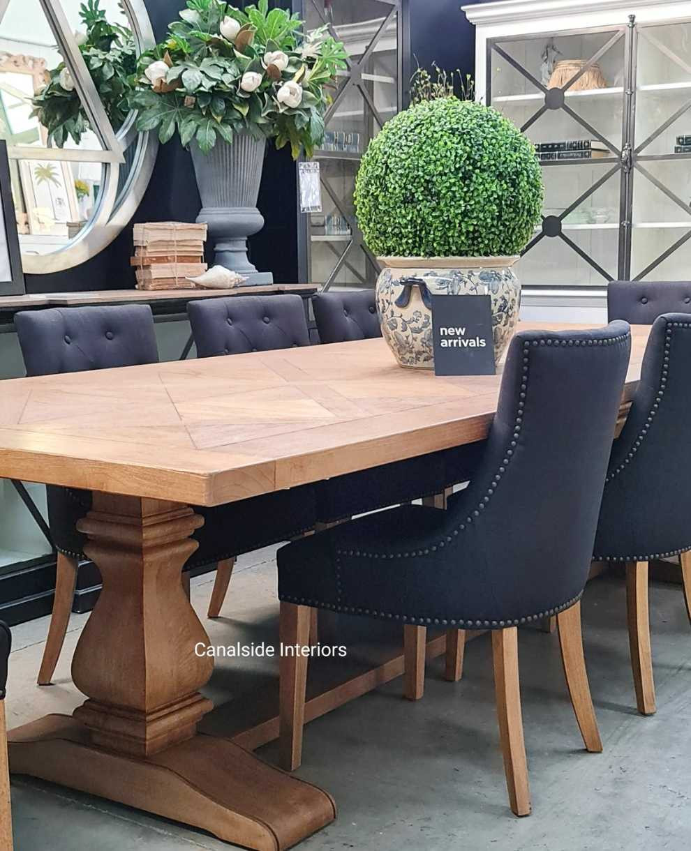 The Hampton Bay Dining Table as the centerpiece in a grand dining room, surrounded by large windows and opulent decor, epitomizing timeless elegance.