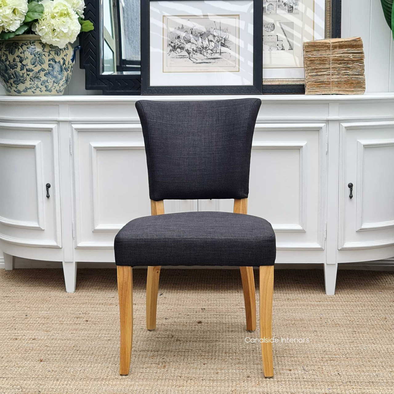 Frontal view of the Mimi Dining Chair in black/charcoal, set against the backdrop of a Hamptons-style dining room, highlighting its distressed natural legs and brass studs.