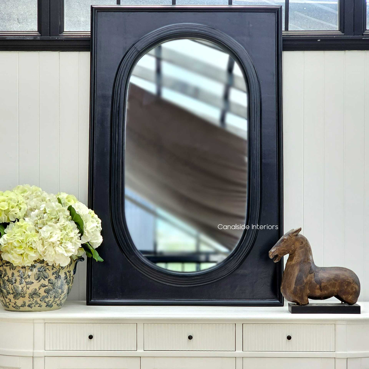 Frontal view of the Ovalis Mirror set against a white paneled wall, adding a classical touch to a modern living space – available at Canalside Interiors.