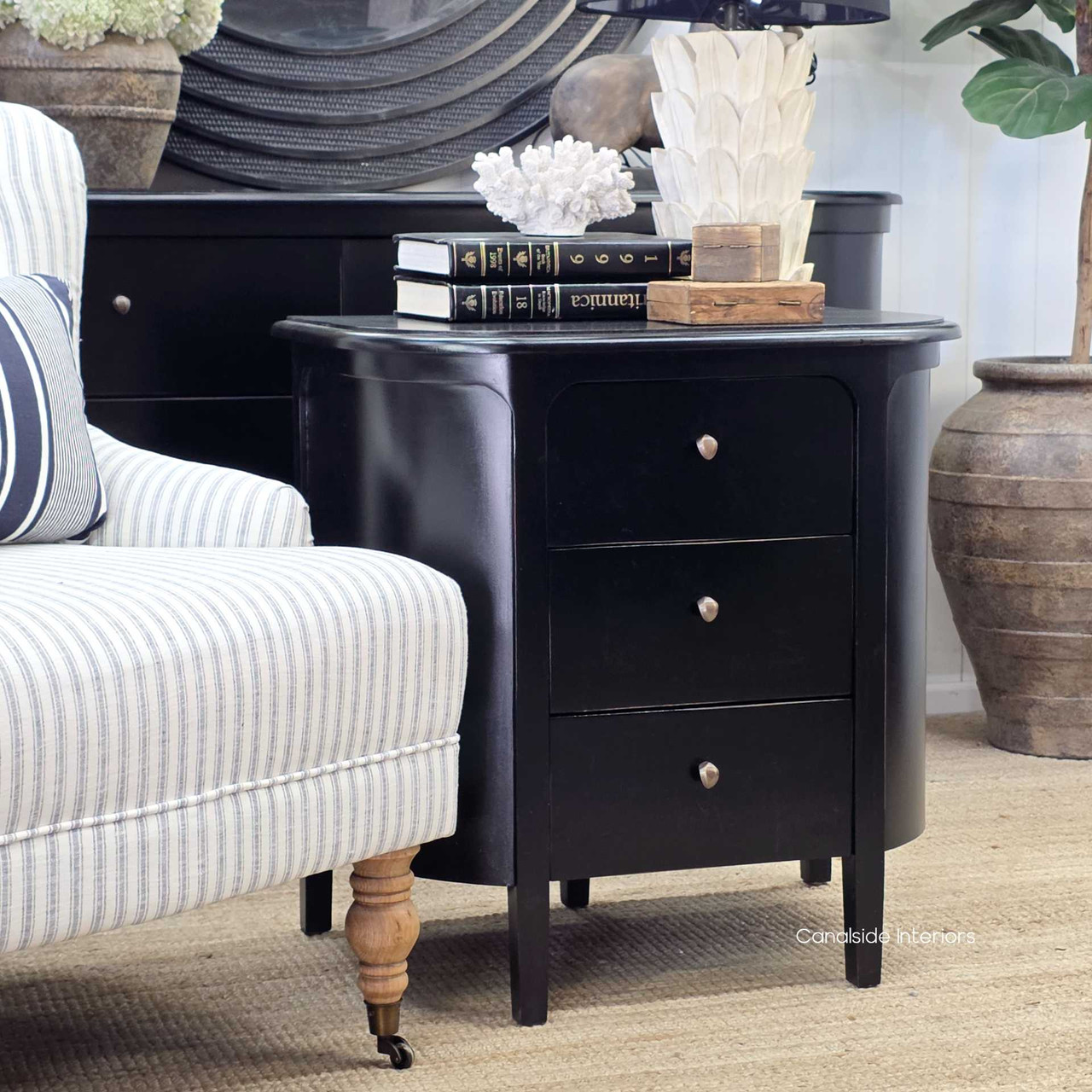 Creemore 3 Drawer Bedside in Black displayed beside an armchair, highlighting its elegant tapered legs and clean-lined profile.