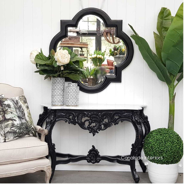 The Baroque Marble Console in Distressed Black stands proudly against a white panel, its Carrara marble top shimmering in Canalside Interiors' elegant display