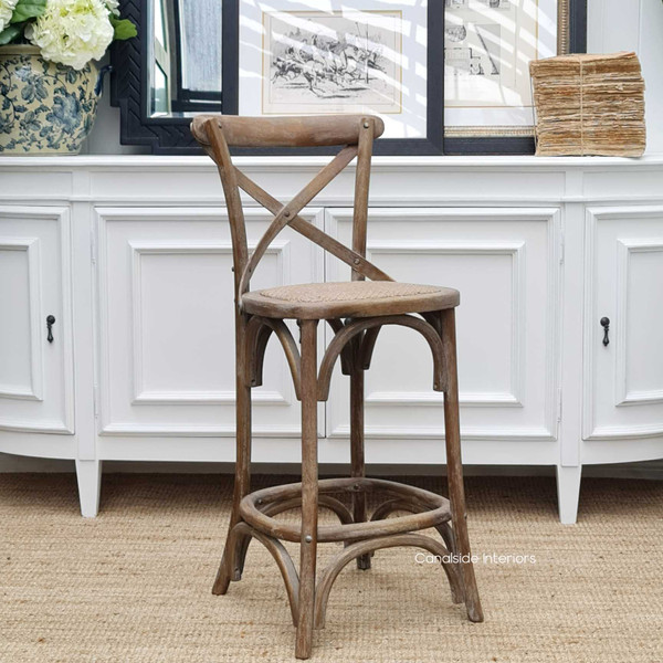 This Hamptons-style Cross Back Kitchen Stool in Distressed Oatmeal stands gracefully against a white paneled backdrop, embodying timeless elegance for your kitchen space