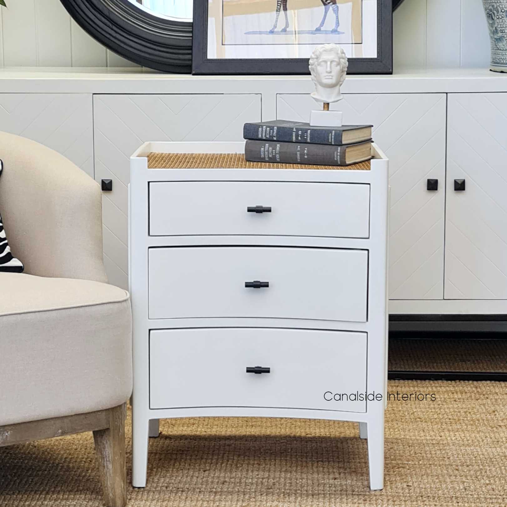 The Tweed Hill Rattan Bedside in distressed white, showcasing its unique concave face and rattan top, set against a neutral bedroom backdrop.