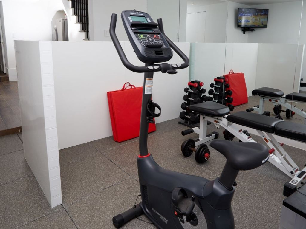 Small home gym with a stationary exercise bike, dumbbells, weight bench, and wall-mounted TV.