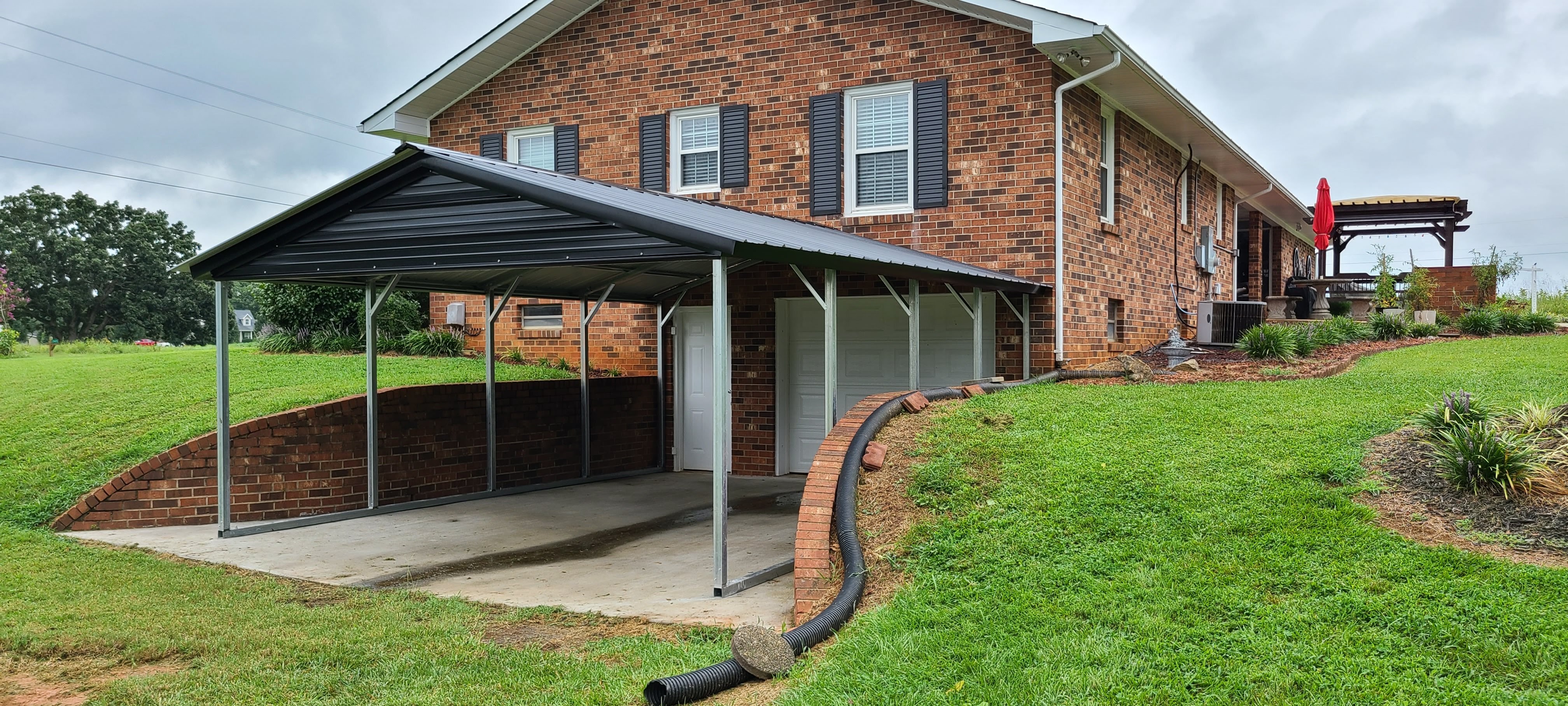 Black metal carport custom-installed on a sloped driveway next to a brick home.