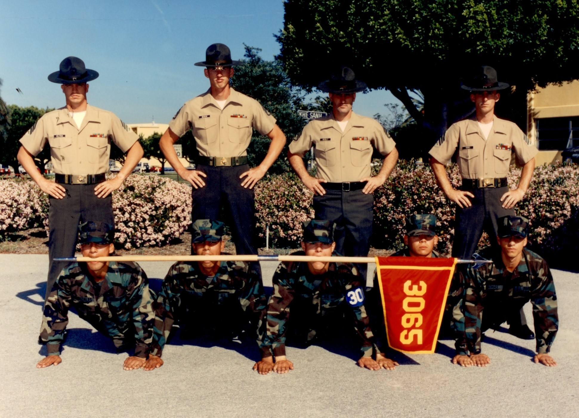 4 drill instructors standing over 4 soldiers holding a flag in their mouths in pushup position