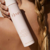 A close-up of a hand holding a bottle of body milk labeled "Fleur's" against a blurred background, featuring a braided hairstyle.