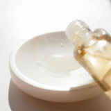 A clear liquid is being poured from a bottle into a small white dish. The surface reflects light softly.