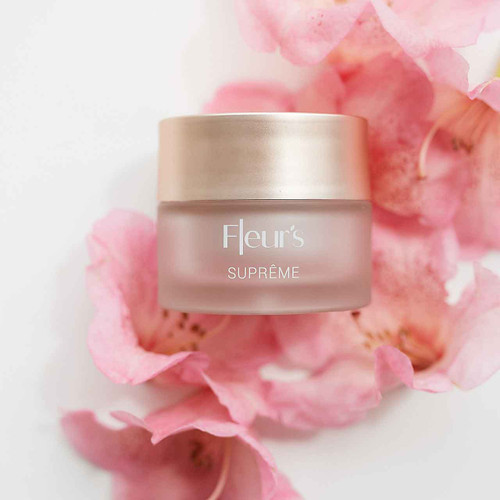A jar of Fleur's Suprême cream surrounded by soft pink flower petals on a light background. A jar of Fleur's Suprême cream surrounded by soft pink flower petals on a light background.