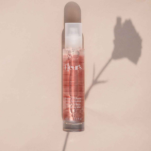 A clear bottle of body exfoliating mist with a pink tint, labeled "Fleur's," casting a shadow of a flower on a beige background. A clear bottle of body exfoliating mist with a pink tint, labeled "Fleur's," casting a shadow of a flower on a beige background.