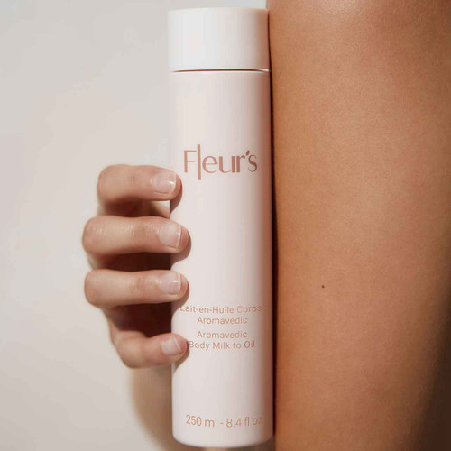 A hand holds a bottle of Fleur's Aromavedic Body Milk to Oil against a light background, showcasing smooth skin. A hand holds a bottle of Fleur's Aromavedic Body Milk to Oil against a light background, showcasing smooth skin.