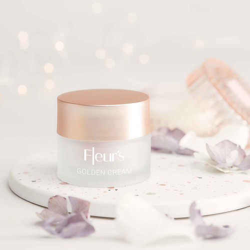 A jar of Fleur's Golden Cream on a speckled surface, surrounded by delicate flower petals and soft lighting.