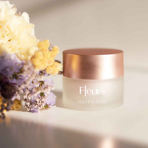 A jar of "Fleur's Happy Skin" cream with a rose gold lid, surrounded by colorful flowers on a light background.