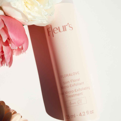 A bottle of Fleur's Floralove Micro-Exfoliant Treatment surrounded by pink and white flowers on a light background.