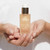 A person holds a bottle of "Fleur's Golden Essence" serum, showcasing its elegant design and label.