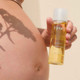 A close-up of a hand holding a bottle of nourishing oil for stretch marks, with a shadow of a plant on a pregnant bare belly.