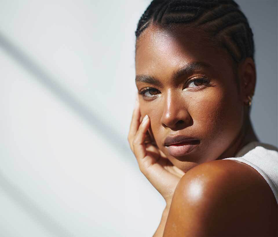 A close-up portrait of a person with braided hair, resting their chin on their hand, illuminated by soft light.