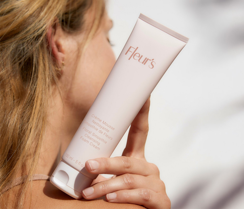 A woman holds a tube of Fleur's Floral Smoothie Cleansing Foam Cream over her shoulder.