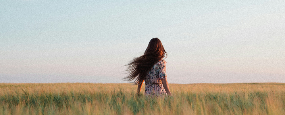 A woman with long hair stands in a field of tall grass, facing away, under a soft sky at dusk.