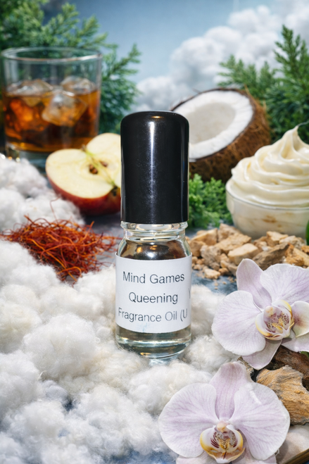 Small glass bottle labeled ‘Mind Games Queening Fragrance Oil (U)’ standing on soft white cotton, surrounded by sliced apple, coconut, vanilla cream, saffron threads, orchids, and a glass of rum, creating a warm, airy amber-woody scene.