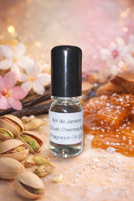 Small perfume oil bottle labeled “Sol de Janeiro Crush Cheirosa 62 Fragrance Oil” centered in a warm, styled scene with pistachios, salted caramel, vanilla beans, and soft jasmine flowers in the background.