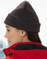 Acrylic beanie in dark grey worn by a person with long hair, paired with a red jacket. The beanie has a logo.