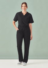 A model wearing unisex black straight leg scrub pants and matching scrub top, standing against a soft green background.