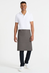 A waist apron in dark grey, featuring a front pocket, worn by a model in a white polo and black pants.