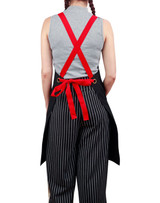 A black apron with red straps worn over a grey top and striped pants, featuring a tie waist and eyelets.
