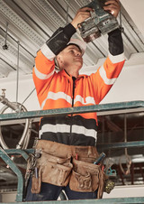 A man in an orange and black hi-vis hoodie uses a tool while standing on a scaffold, wearing a tool belt.