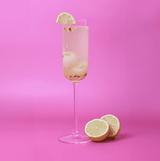 A ribbed, clear glass champagne flute filled with a light beverage, garnished with a lemon slice, against a pink background.
