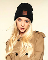 A black knitted beanie with a pom pom, featuring a branded patch, modelled by a person with long blonde hair.