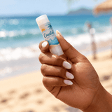 A hand holding a white tube of Twist Lip Balm with a beach and ocean background. The product features a logo.