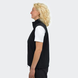 A woman stands sideways wearing a black vest with a high collar. The vest is sleeveless and fitted.