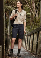 A woman in a beige long sleeve button-up shirt and navy shorts stands on a forest walkway, carrying a bag.