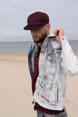 A man at the beach wears a burgundy cap and a denim jacket with graphic details. The ocean is in the background.
