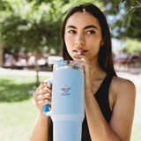 A drink bottle in light blue with a handle, featuring a logo and a clear lid, held by a person outdoors. A drink bottle in light blue with a handle, featuring a logo and a clear lid, held by a person outdoors.
